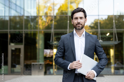 Confident businessman standing outdoors holding a laptop, representing modern work, technology, success, and corporate professionalism in front of a glass office building