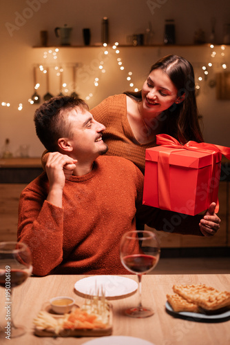 Happy couple in love celebrating romantic Valentine Day, woman holding gift box, hugging kissing her husband during festive dinner at home