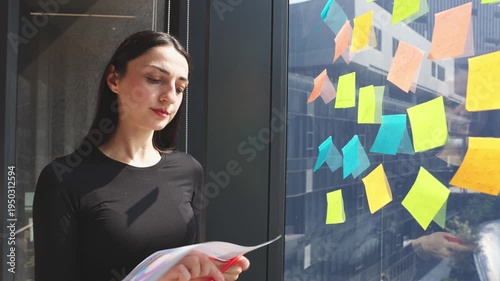 Young professional organizing ideas on colorful sticky notes while reviewing business chart near office window during planning session.