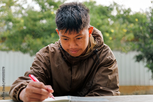 Asian teenage boy with concentrating on writing homework in notebook outdoors