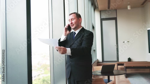 Professional businessman speaking on smartphone while holding financial report near office window during work discussion.