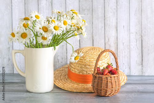 Summer still life; wicker basket with ripe strawberries, bunch of white daisy flowers in white jug and straw hat on a wooden background