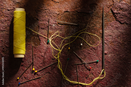 Macro photo of sewing tools on rusty metal surface — yellow thread spool, needles and pins. Professional still life photography for design and decoration