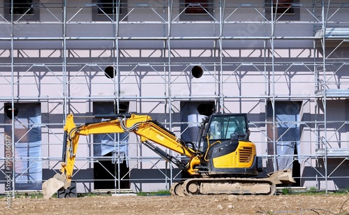 Crawler compact excavator in a dirt area next to a building under construction surrounded by scaffolding. Side view.