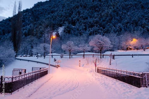 A peaceful winter scene at twilight featuring a snow-covered pedestrian path with visible footprints and tire tracks. 