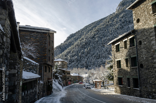 A ground-level perspective of a narrow road winding through an alpine village. The scene features traditional multi-story buildings made of local dark stone and slate