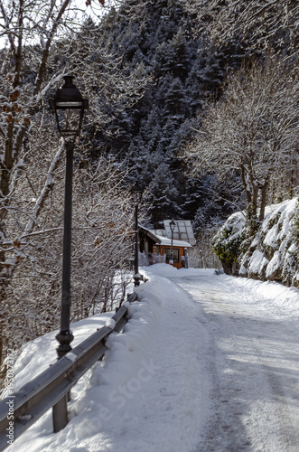 Snow-covered mountain path lined with vintage street lamps and trees, leading to a small wooden cabin. A peaceful winter scene surrounded by forest and alpine landscape.