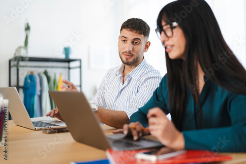 A man and a woman collaborate on laptops at a desk, discussing ideas and working together on a project in a bright office environment.