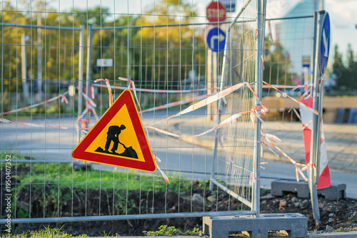Road construction warning sign, metal fence blocking street, barrier tape, temporary fencing around excavation on urban roadway. Road construction safety warning, traffic restriction, street repair