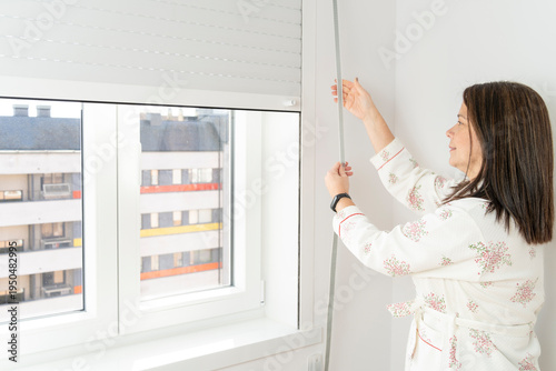 Foto Woman in bathrobe pulling cord to raise roller shutter in home