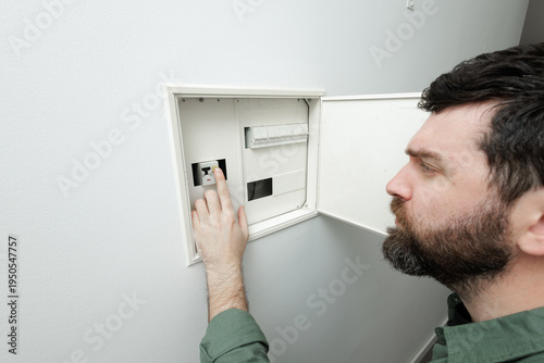A man with a beard inspects a home electrical fuse box to reset a tripped breaker or test the power. Stock photo