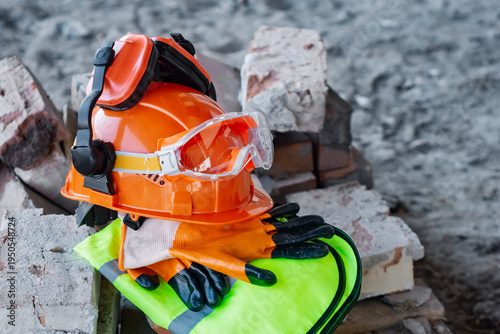 Orange hard hat with ear muffs and clear safety goggles with work gloves and a neon green high-visibility safety vest with reflective stripes. Concept on a stack of bricks at a construction site.