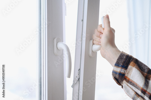 woman hand opening white plastic window by handle, in house or apartment or office, young caucasian girl in warm plaid shirt