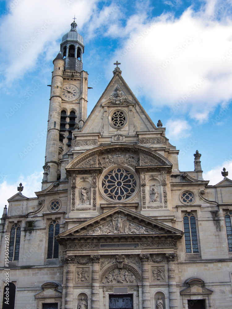 Fototapeta premium Dramatic low-angle view of the highly detailed facade and distinctive clock tower of the historic Church of Saint-Étienne-du-Mont under a vibrant, cloudy blue sky.