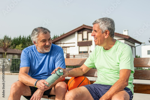 Two cheerful elderly men friends relaxing on a bench after playing basketball, one passing a water bottle to the other, embodying active aging and shared wellbeing