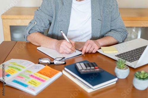 Woman writing in planner while managing finances at a wooden desk with laptop, calculator, sticky notes and receipts — focused on budgeting, business planning and organization