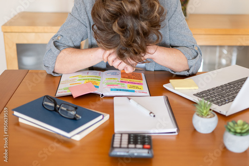 Woman feeling overwhelmed and stressed, resting her head on folders and a color-coded planner filled with tasks, finances, and budget details, indicating burnout and exhaustion from corporate work
