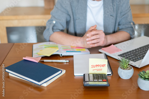 Business woman sitting at a messy desk with a laptop, calculator, and sticky note reading support while planning finances, operations, and seeking solutions for corporate budget and tax issues