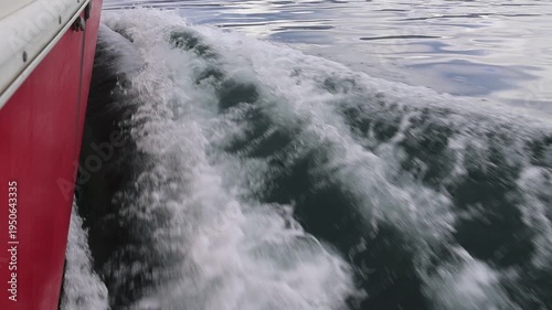 The red hull of a small boat is visible on the left side of the video, while on the right side, water forms ripples and foam due to the boat's movement.