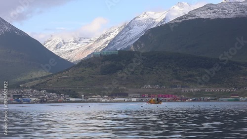 A scene filmed from a boat showing a landscape involving a small vessel sailing on the Beagle Channel, the port city of Ushuaia in the background, and the imposing snow-capped mountains behind the cit