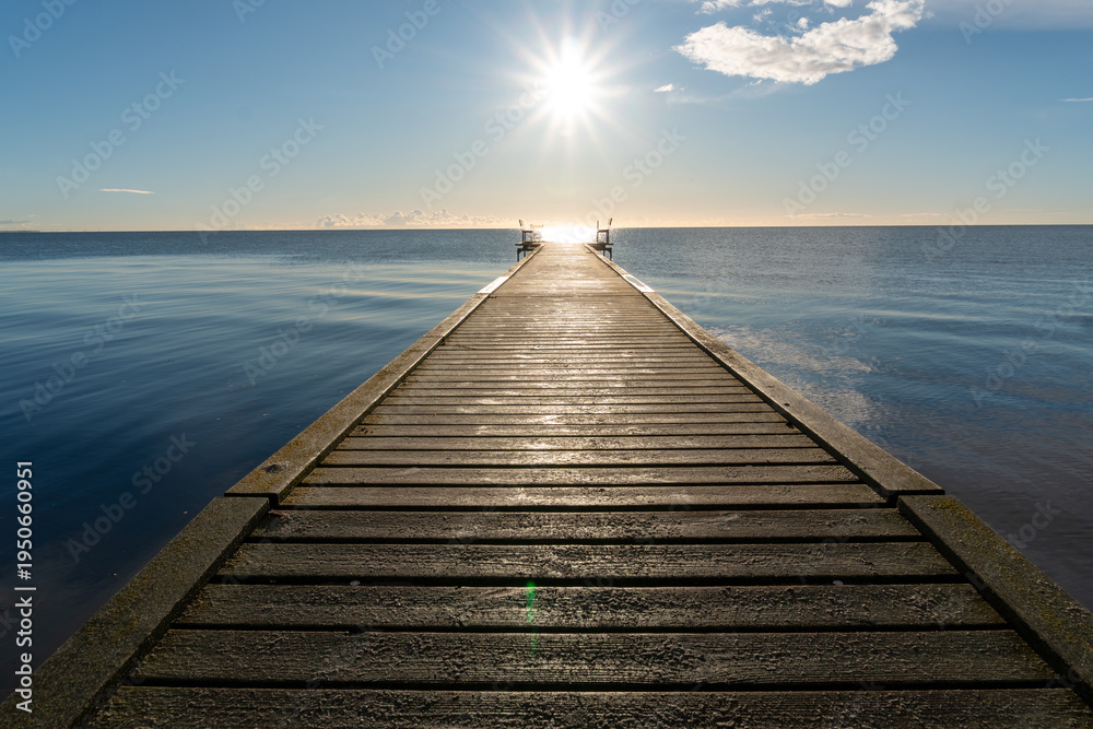 Obraz premium Straight wooden jetty extending toward the open sea with bright sun above the horizon. The calm water reflects the sky creating a peaceful marine landscape often associated with summer travel, relaxat