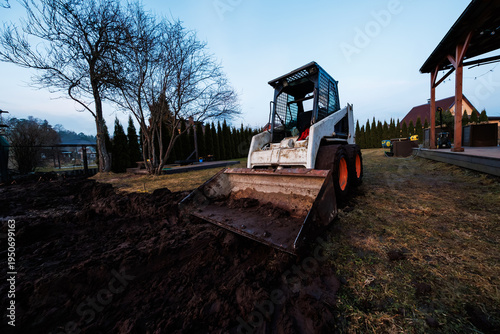 Skid steer grading wet soil in residential yard with arborvitae