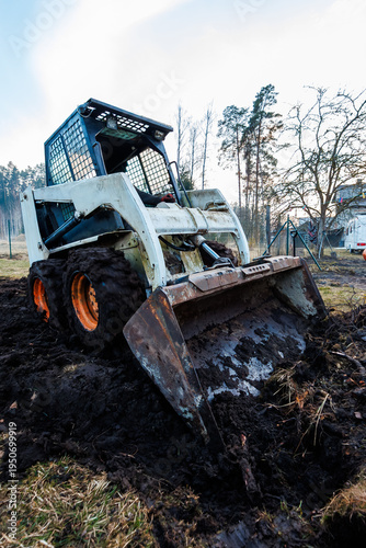 Skid Steer Loader Digs Wet Soil in Rural Yard with Swing Set