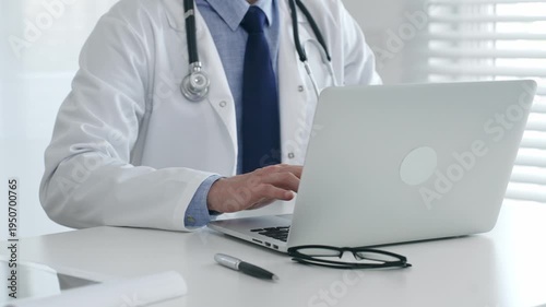 Male doctor working on laptop, wearing lab coat, stethoscope, eyeglasses in modern clinical workspace. Medicine and health care concept