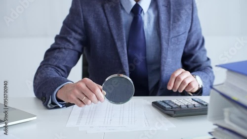 Businessman or accountant in a blue suit and tie sitting at a desk, using a magnifying glass to analyze financial documents with a calculator nearby, close up. Audit and taxes theme in business