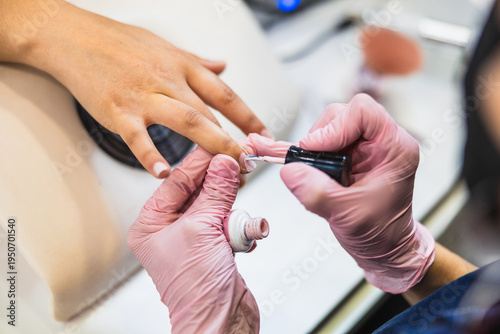 Professional nail technician wearing pink gloves carefully applying light pink nail polish on customer's fingernails in a beauty salon