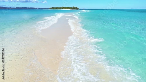 Aerial drone view above a long white sandbar surrounded by crystal clear turquoise ocean water, leading toward a small tropical island in Madagascar