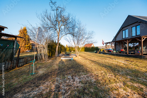 Late afternoon light casts long shadows on a modern dark clad house with covered deck, large windows, arborvitae, leafless trees, picnic table, toys, and lawn tractor.