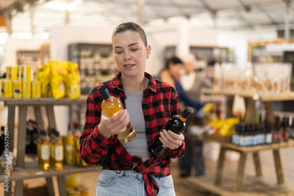 Obraz premium Girl chooses semi-sweet wine while standing near the shelves with alcohol in a hypermarket. European chooses bottles of wine in a store against the background of customers