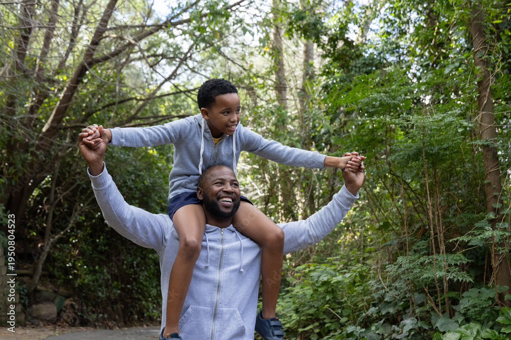Obraz premium African American father and son walking wooded path, carrying son on shoulders in gray hoodies