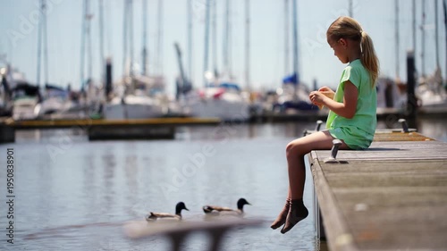 A young blonde girl in a green outfit sits on a wooden dock, dangling her feet. She throws food to ducks swimming in the marina, with sailboats blurred in the background on a sunny day.