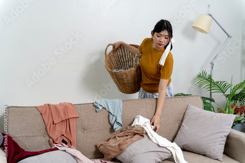 Obraz na płótnie Asian Woman Collecting Scattered Clothes From Sofa Into Wicker Laundry Basket Li