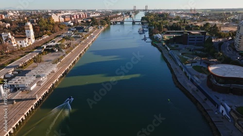 Aerial view shows a long river in Spain with a pier, a modern complex, twin lift bridge towers, palm trees, a golden domed tower, a marina, rowers, and a motorboat.