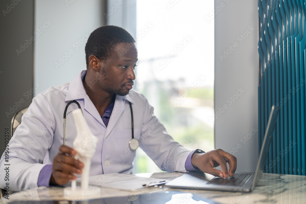 Fototapeta premium African American male physical therapist doctor looking at laptop holding anatomical knee joint model in rehabilitation center, Orthopedic medical clinic, healthcare and physiotherapy concept
