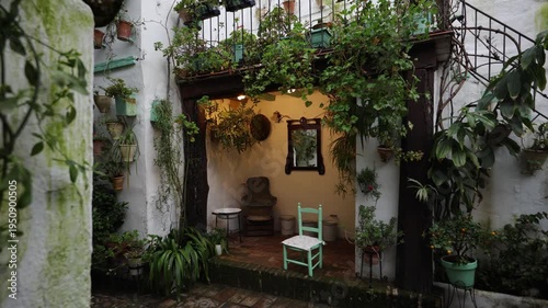 Whitewashed courtyard in Spain with wrought iron balcony, wall pots, turquoise chair, warm lamp, ferns, mirror, orange tree, vines, and spiral staircase.