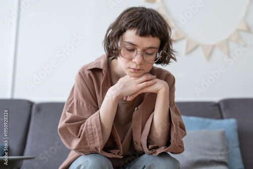 Sad upset teenage girl sitting on sofa, looking down with withdrawn expression, resting chin on hands. Teen uncertainty, social isolation, low self-confidence, introversion and emotional discomfort.
