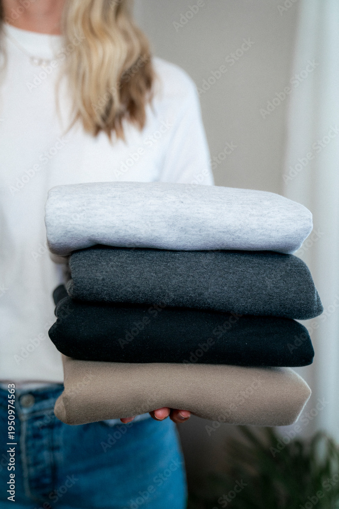 Fototapeta premium Vertical lifestyle image of blonde woman in jeans and white shirt holding neutral sweatshirts in clean bedroom, small business owner or laundry concept