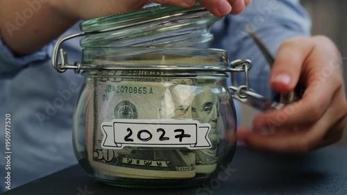 Female hands take money from Glass jar full of American currency dollars cash banknote with text 2027 year. Preparation saving money. Moderate consumption and economy. Collecting money