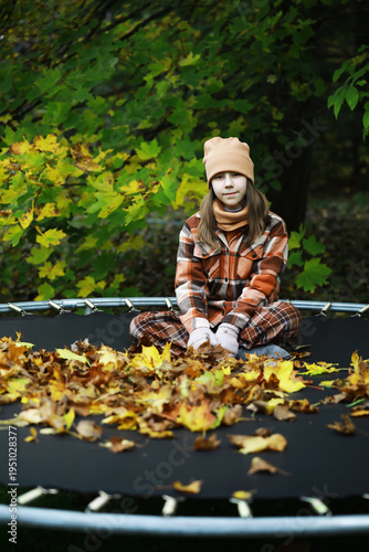 Foto Young Girl in Autumn Attire Sits on Trampoline Amidst Colorful Fallen Leaves in