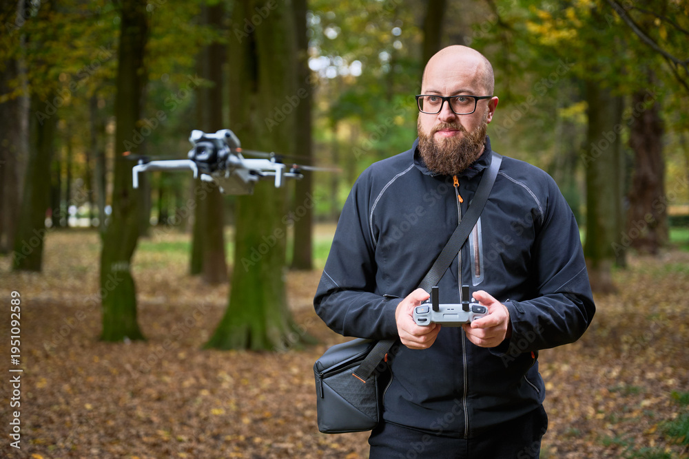 Fototapeta premium In a peaceful forest during autumn, a man with a beard expertly controls a drone, focused on its flight. Leaves cover the ground, creating a vibrant atmosphere.