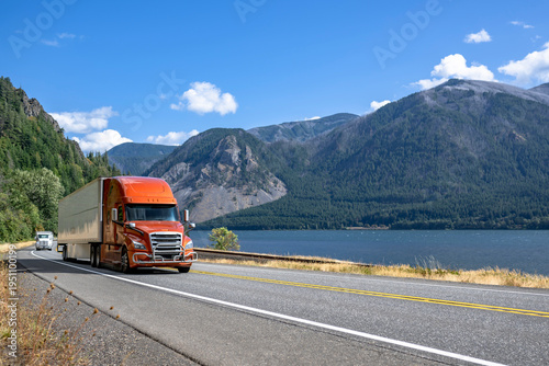 Orange big rig semi truck with grille guard transporting cargo in dry van semi trailer running on the highway road along the Columbia River in Columbia River Gorge area
