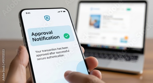 A hand holds a smartphone displaying an approval notification for a transaction after successful secure authentication, with a laptop in the background.