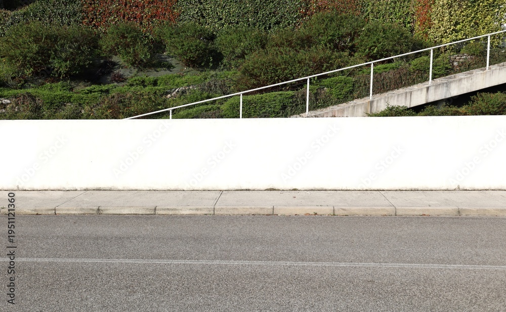 Fototapeta premium White plaster fence with a staircase, vegetation and multicolored hedge on behind. Concrete sidewalk and street in front. Background for copy space.