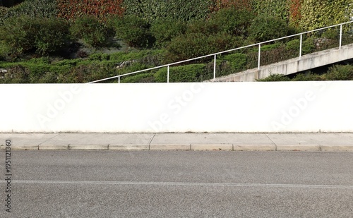 White plaster fence with a staircase, vegetation and multicolored hedge on behind. Concrete sidewalk and street in front. Background for copy space.