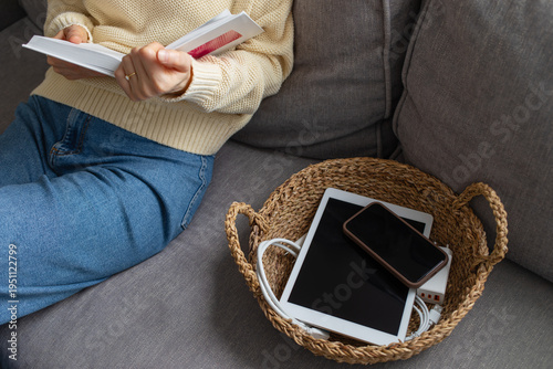 Digital detox concept from woman reading paper book while phone and tablet gadgets lying offline in basket. Home and mental leisure instead social media and technology addiction.