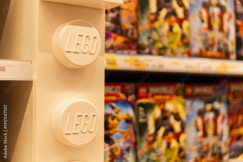 Naklejka premium Chemnitz, Germany - March 14, 2026: A shelf features a large LEGO block with the brand name. Behind it, various LEGO sets are arranged neatly. Customers browse the selection in a retail store
